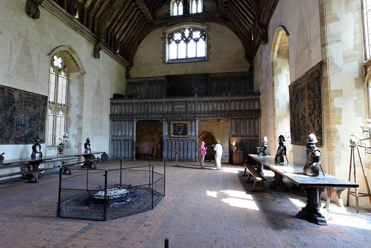 Great Halls and Medieval Castle Life 1 The interior of a medieval great hall with a high open timber roof: an octagonal open hearth sits in the middle of the tiled floor inside a modern mesh guard, long trestle tables with pieces of armour on them run down both side walls beneath tapestries and traceried windows, and a carved timber screen with a gallery above it closes the far end.
