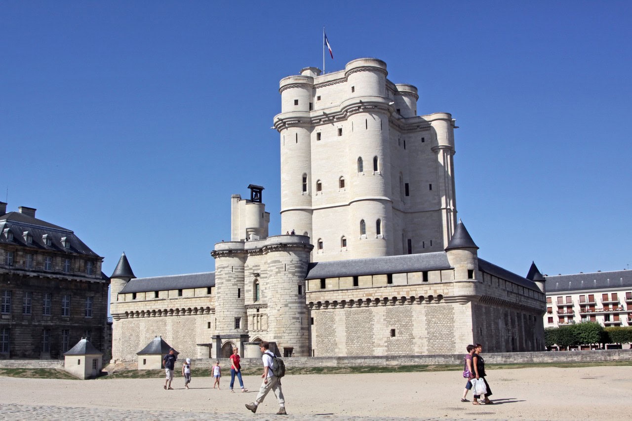 The tall pale stone donjon of the Château de Vincennes rising behind the crenellated wall of its rectangular chemise, with a gate tower at the foot of the great tower and visitors crossing the open esplanade in front.