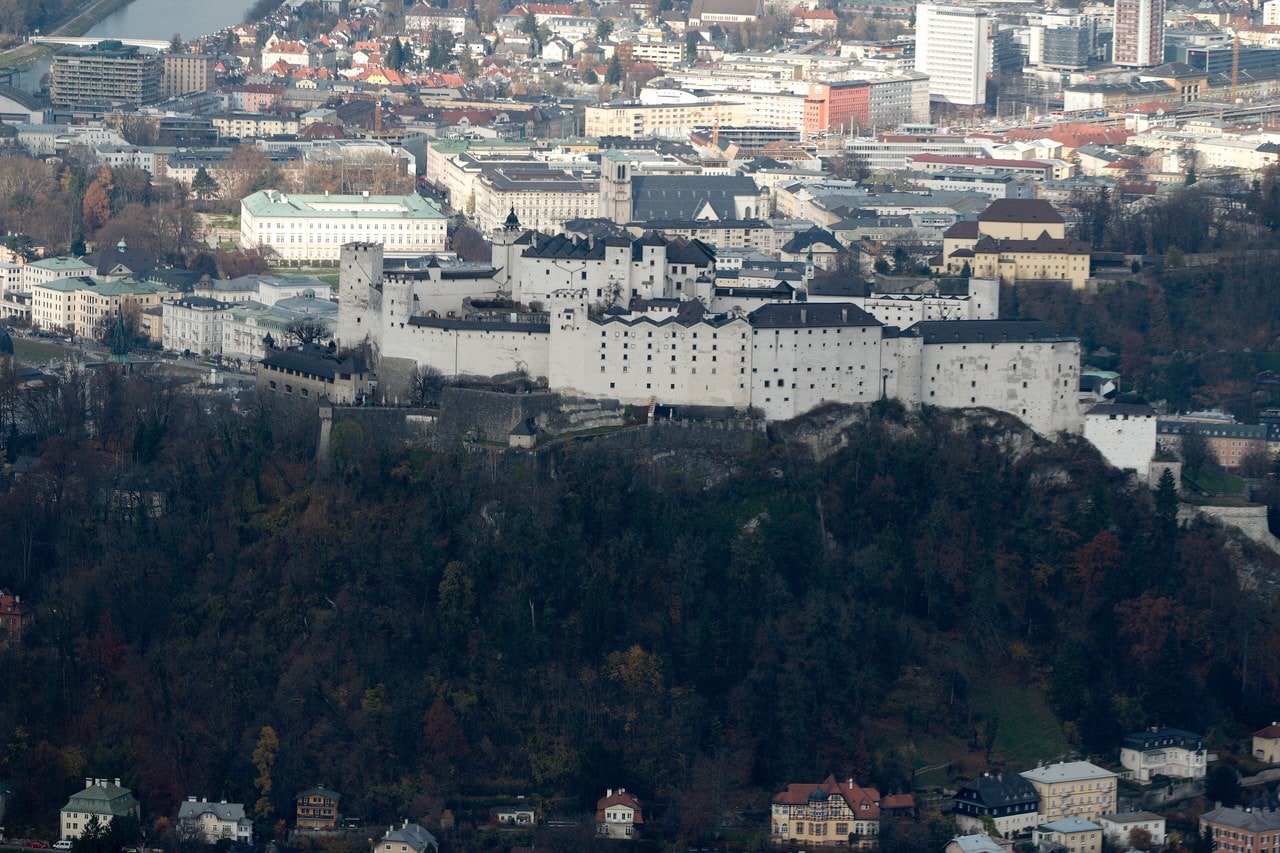 Hohensalzburg reads as a long band of white rendered masonry along the top of its wooded rock, with the roofs of Salzburg's old town and the modern city spread out beyond it. Hohensalzburg reads as a long band of white rendered masonry along the top of its wooded rock, with the roofs of Salzburg's old town and the modern city spread out beyond it.