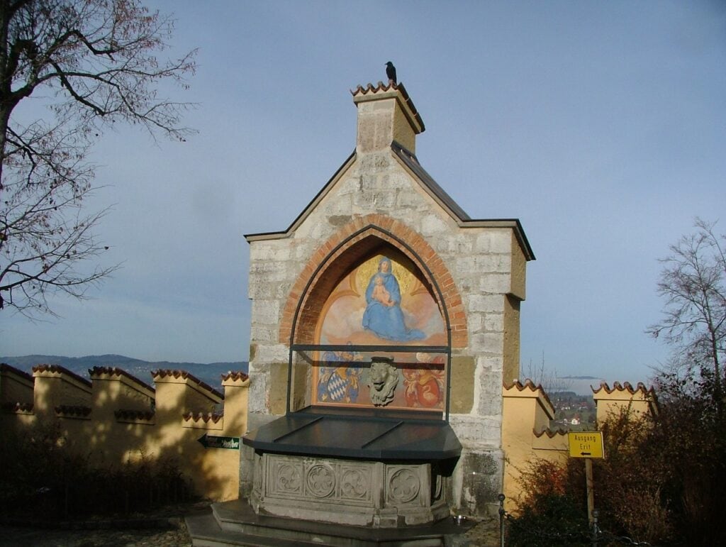 Hohenschwangau Castle 9 The Marienbrunnen at Hohenschwangau, a gabled wall fountain set in the crenellated Wehrmauer, a carved lion's-head spout between the Bavarian lozenge and the Schwangau arms below a painted Madonna and Child, above a stone basin