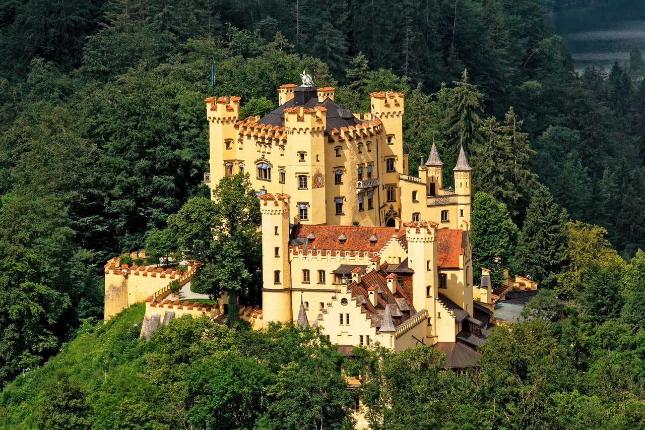 Hohenschwangau Castle seen from above, an ochre block with crenellated parapets and red roofs on a wooded hill, the castle wall running below it Hohenschwangau Castle seen from above, an ochre block with crenellated parapets and red roofs on a wooded hill, the castle wall running below it
