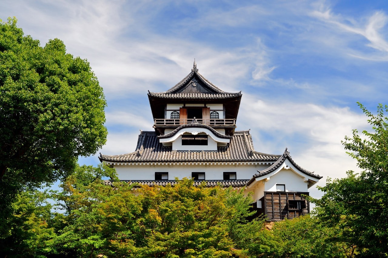 The multi-story keep of Inuyama Castle rising above green trees against a blue sky, its walls white-plastered under dark curving tiled roofs, with an undulating cusped gable on the face below the top floor and a railed open veranda running around the topmost story. The multi-story keep of Inuyama Castle rising above green trees against a blue sky, its walls white-plastered under dark curving tiled roofs, with an undulating cusped gable on the face below the top floor and a railed open veranda running around the topmost story.