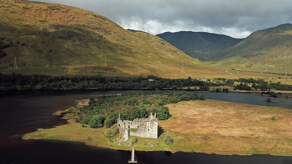 Kilchurn Castle 2 Aerial view of Kilchurn Castle at the end of a low green peninsula running out into the dark water of Loch Awe, with a glen and hills rising behind.