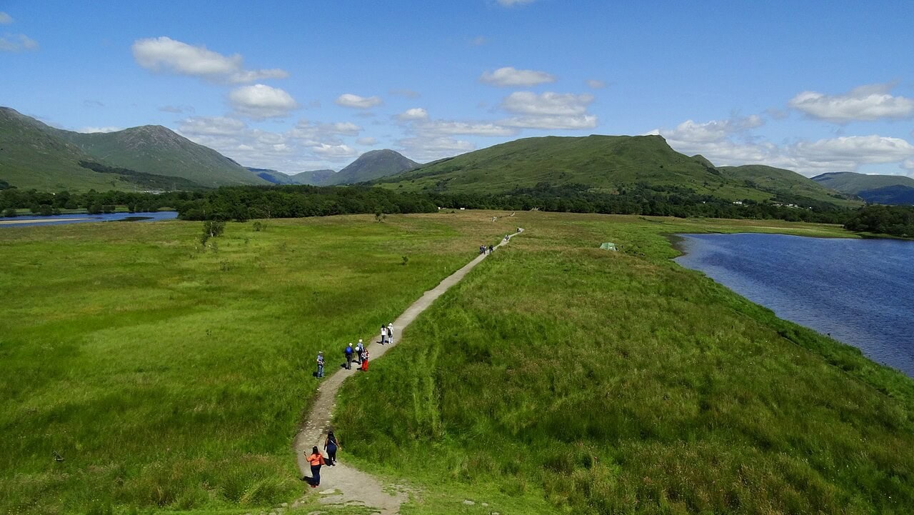 Walkers strung out along a narrow track running on a low raised line across flat grazing ground at Kilchurn Castle, with water lying on both sides and green hills beyond. Walkers strung out along a narrow track running on a low raised line across flat grazing ground at Kilchurn Castle, with water lying on both sides and green hills beyond.