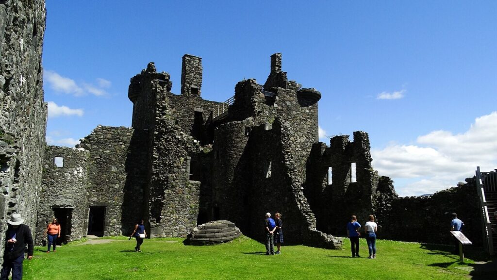 Kilchurn Castle 3 People standing on the grass of Kilchurn Castle's courtyard under a blue sky, with the roofless tower house and its ranges behind and the fallen stone turret on the ground nearby.