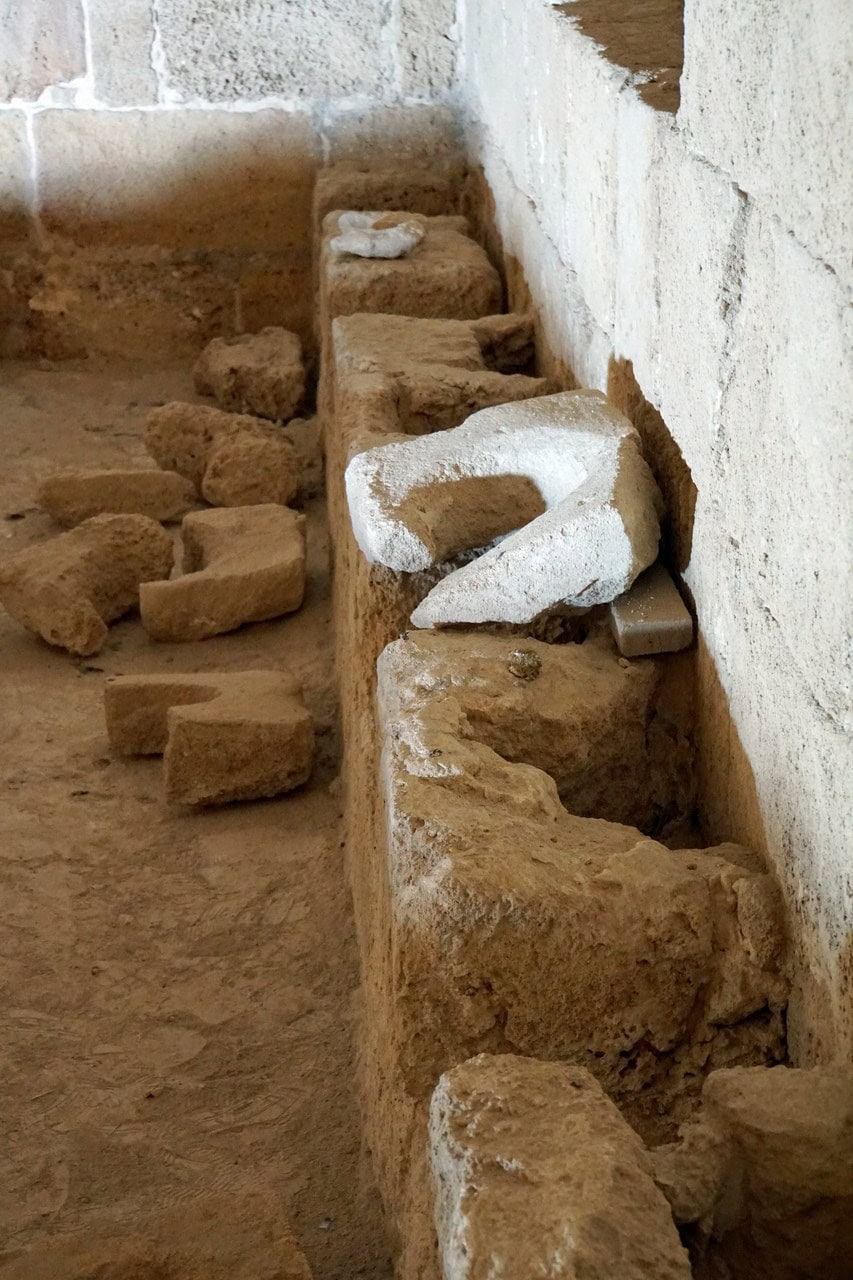 A long excavated trench beside a plastered wall, with a row of shaped stone blocks set along its edge and further loose blocks lying in the cut. A long excavated trench beside a plastered wall, with a row of shaped stone blocks set along its edge and further loose blocks lying in the cut.
