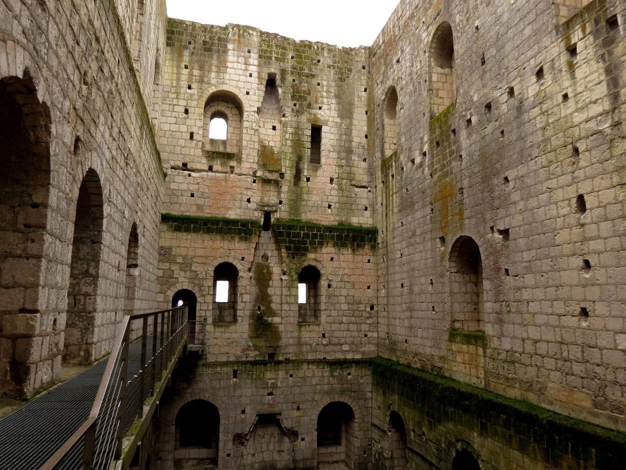 The hollow interior of the great tower at Loches, open to the sky, with tiers of arched window embrasures, rows of square joist sockets cut into the masonry, tapering fireplace flues on the far wall, and a modern steel walkway at the left.