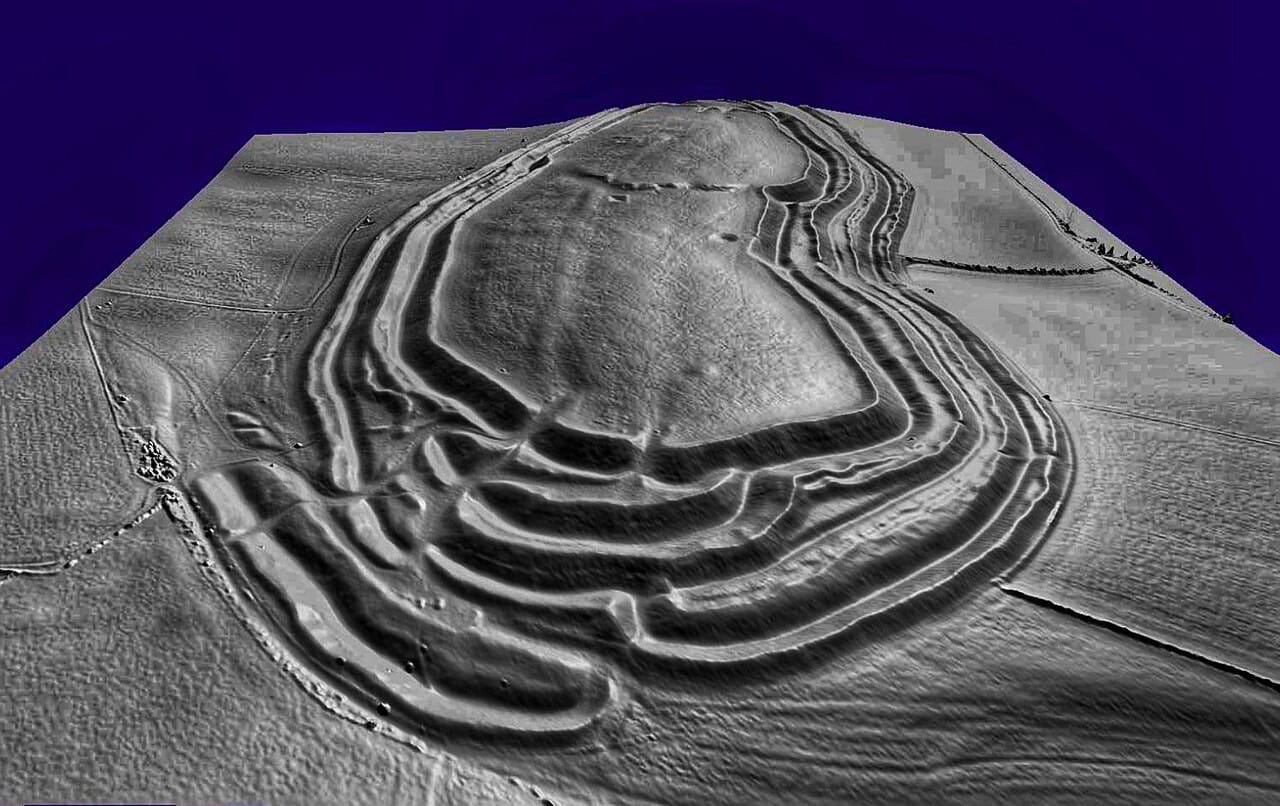 A grey shaded-relief terrain model of a hillfort against a dark blue background, the vegetation stripped away so that multiple concentric ramparts and ditches show as parallel ridges wrapped around an elongated hilltop, with complex entrance earthworks at the near end. A grey shaded-relief terrain model of a hillfort against a dark blue background, the vegetation stripped away so that multiple concentric ramparts and ditches show as parallel ridges wrapped around an elongated hilltop, with complex entrance earthworks at the near end.
