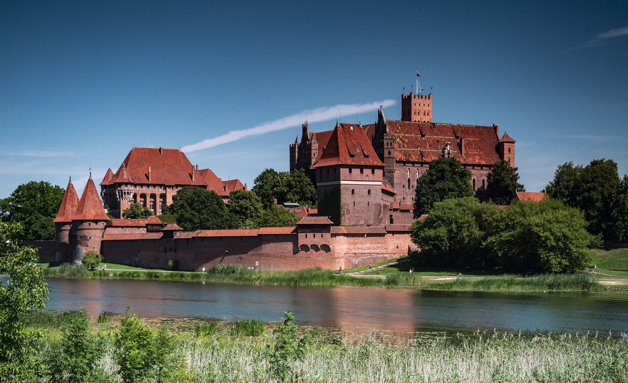 Malbork Castle seen from across the Nogat, built in red brick from the waterline to the roof ridge, with a round gate tower at the left and the tall block of the High Castle at the right. Malbork Castle seen from across the Nogat, built in red brick from the waterline to the roof ridge, with a round gate tower at the left and the tall block of the High Castle at the right.