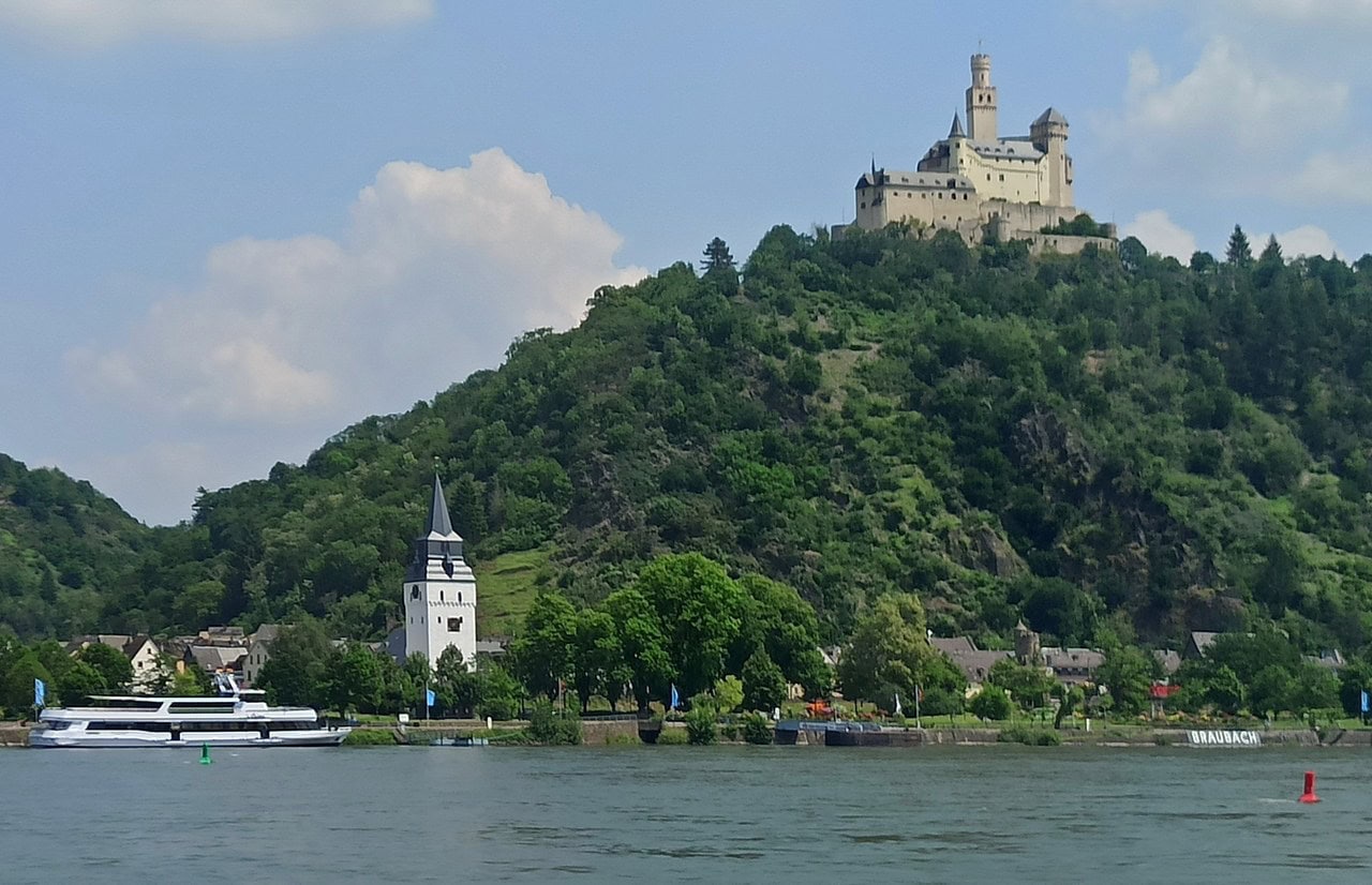 Marksburg Castle on the crest of a steep wooded hill above the Rhine, with the riverside town of Braubach and its white church tower along the bank below and a passenger boat on the water. Marksburg Castle on the crest of a steep wooded hill above the Rhine, with the riverside town of Braubach and its white church tower along the bank below and a passenger boat on the water.