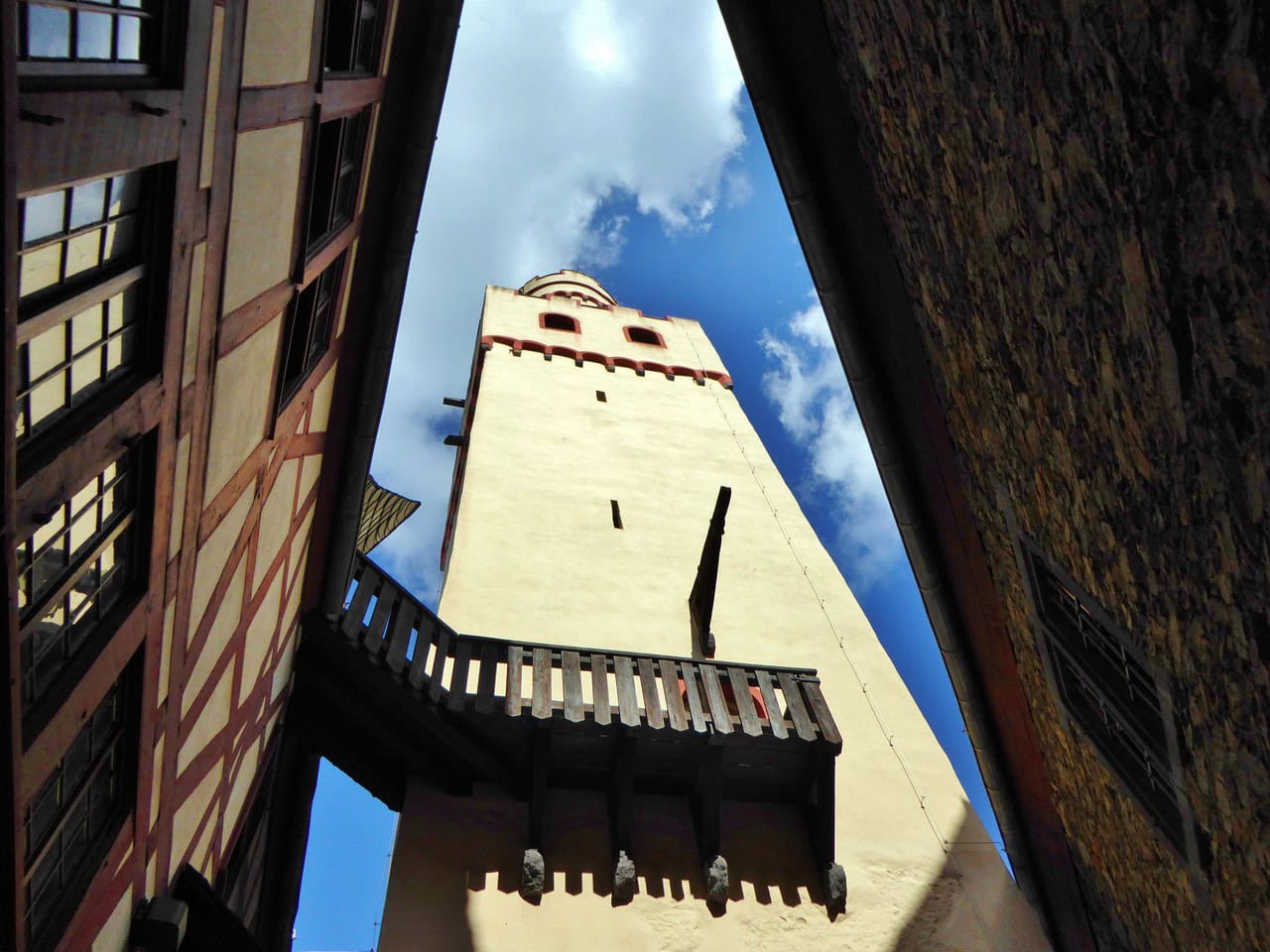 Looking steeply up at the Bergfried of Marksburg from a narrow courtyard between a timber-framed range and a stone wall, showing the tall square white-rendered shaft, a corbelled frieze and battlements, and the round cap on top.