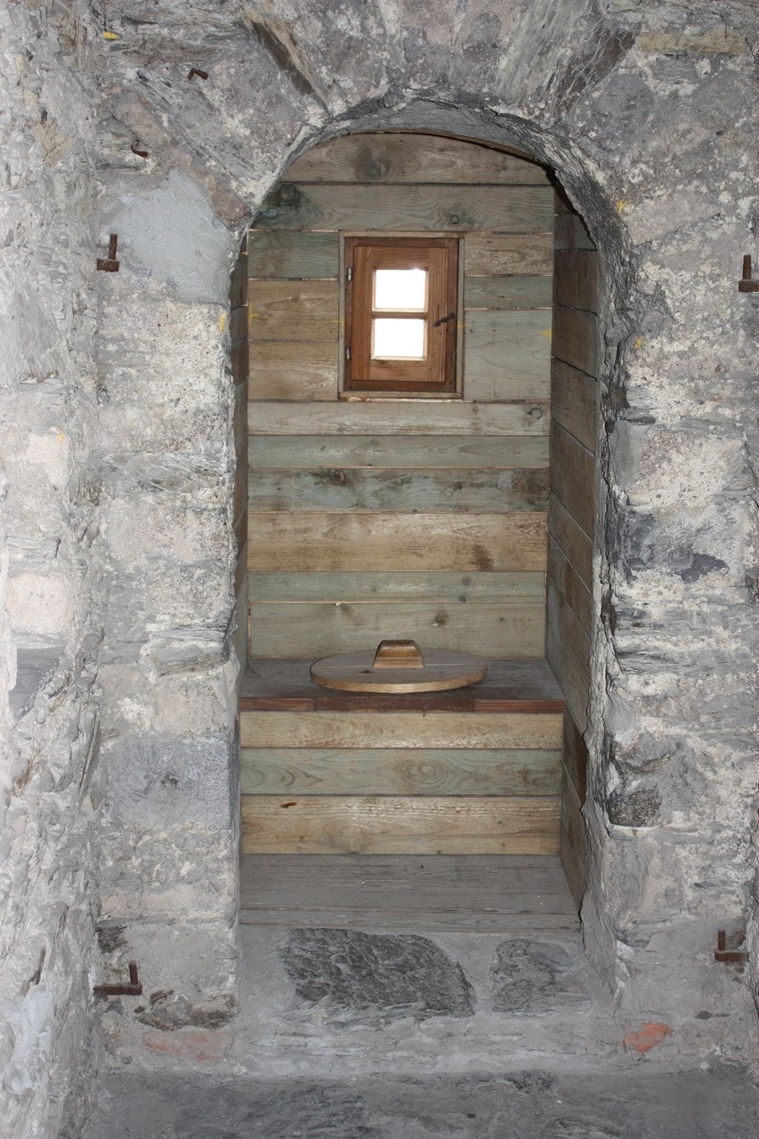 A narrow closet lined with pale timber boards seen through a rough stone arch, lit by a small square window, with a round wooden lid set into a bench seat. A narrow closet lined with pale timber boards seen through a rough stone arch, lit by a small square window, with a round wooden lid set into a bench seat.