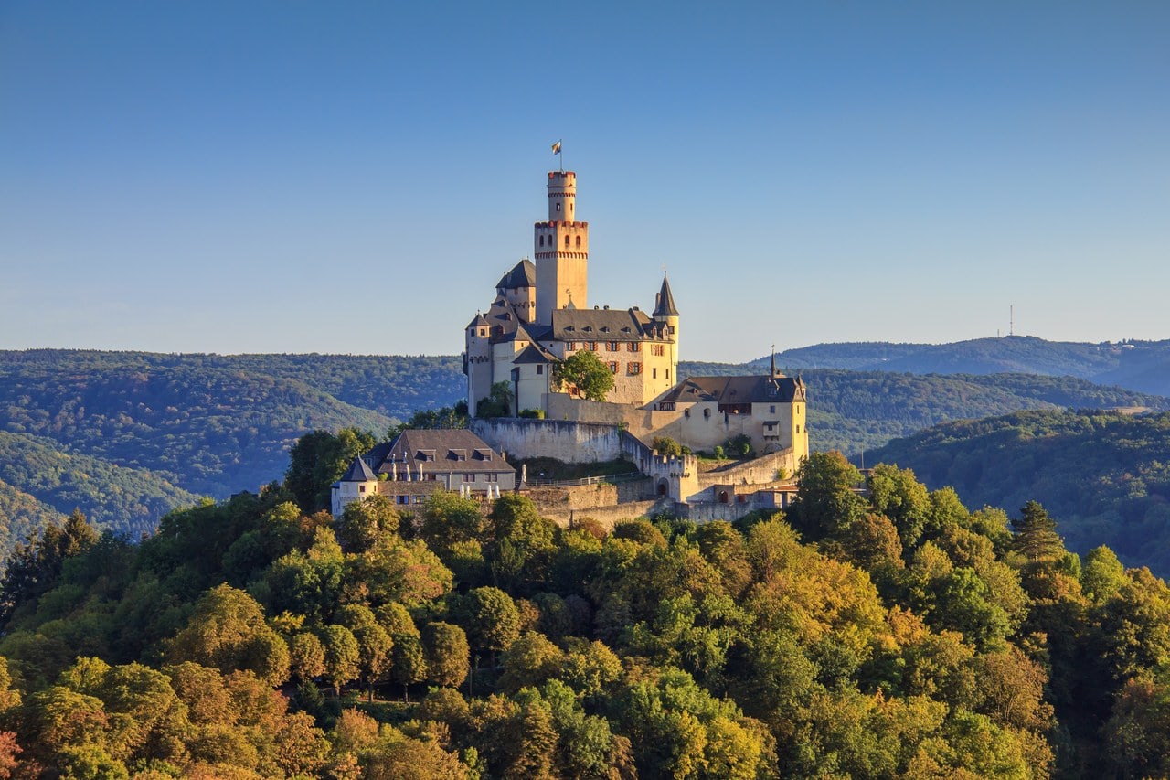 Marksburg standing on the crown of a steep wooded hill in low sunlight, its pale keep with a red-banded battlemented top rising above slate-roofed ranges, with wooded ridges receding into haze behind. Marksburg standing on the crown of a steep wooded hill in low sunlight, its pale keep with a red-banded battlemented top rising above slate-roofed ranges, with wooded ridges receding into haze behind.