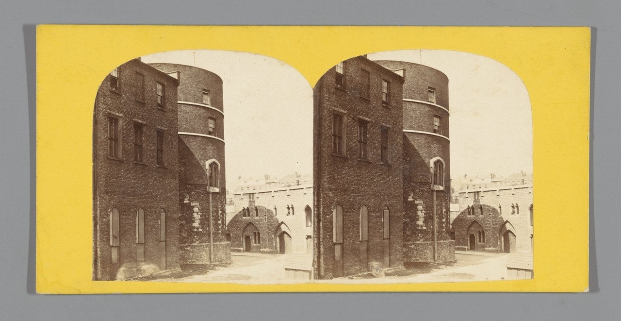 A yellow-mounted stereograph card carrying two arched photographs of the round Martin Tower at the Tower of London with a tall dwelling range built against it. A yellow-mounted stereograph card carrying two arched photographs of the round Martin Tower at the Tower of London with a tall dwelling range built against it.