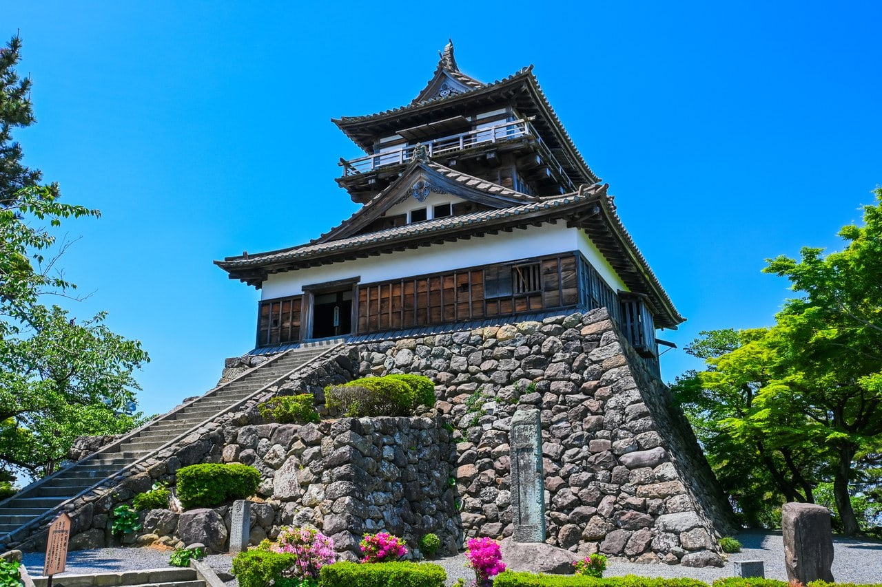 The timber keep of Maruoka Castle in Sakai, Fukui, a tower of roofed stages with white plastered and boarded walls, standing on a battered base of rough uncut stone, a steep flight of stone steps climbing to the entrance and a railed veranda around the top story, under a deep blue sky. The timber keep of Maruoka Castle in Sakai, Fukui, a tower of roofed stages with white plastered and boarded walls, standing on a battered base of rough uncut stone, a steep flight of stone steps climbing to the entrance and a railed veranda around the top story, under a deep blue sky.