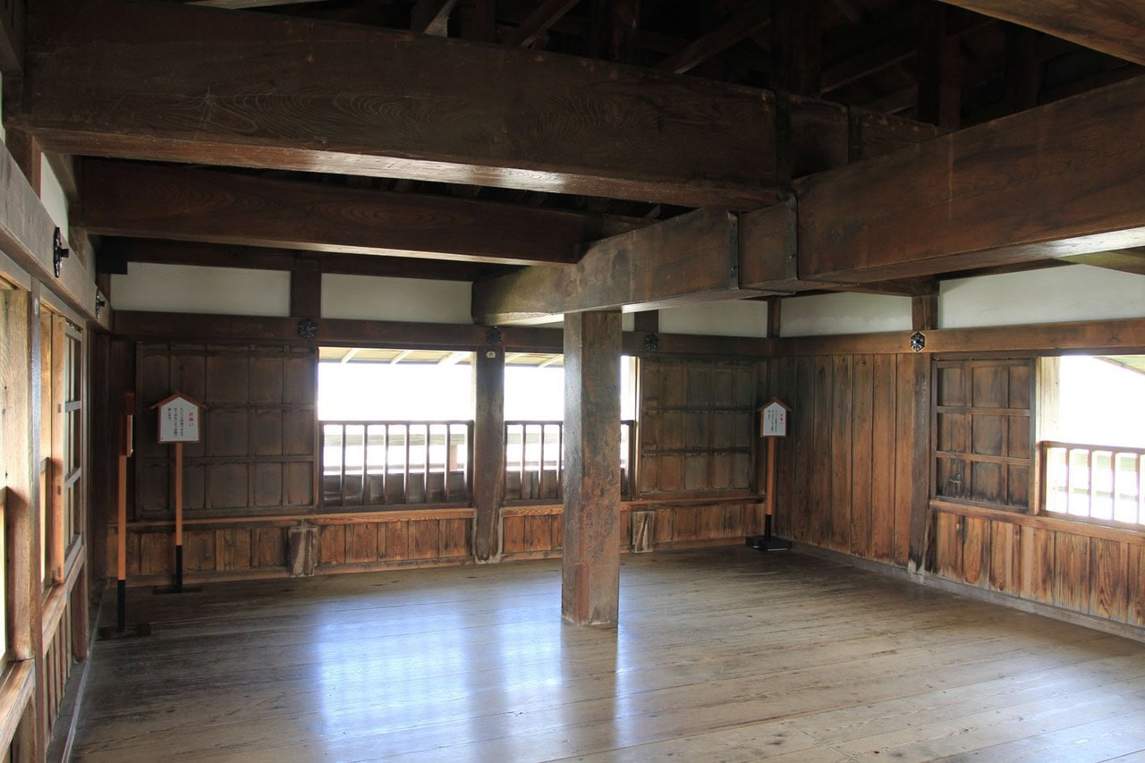 The interior of a story of the Maruoka Castle keep, with a bare boarded floor, a heavy squared timber post rising to deep sawn crossbeams, plank-panelled walls and low railed openings looking out under the eaves. The interior of a story of the Maruoka Castle keep, with a bare boarded floor, a heavy squared timber post rising to deep sawn crossbeams, plank-panelled walls and low railed openings looking out under the eaves.