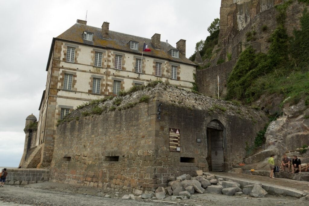 Mont-Saint-Michel 3 A battered stone wall with a round corner bartizan and low openings, a slate-roofed building standing above it, the strand in front