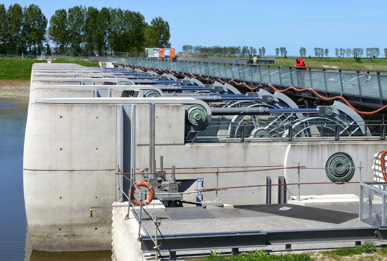 A concrete dam carrying a row of hydraulic gate machinery and a walkway, with water either side A concrete dam carrying a row of hydraulic gate machinery and a walkway, with water either side