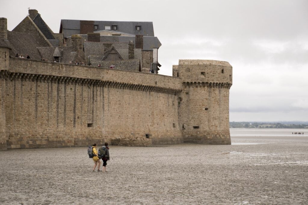 Mont-Saint-Michel 9 A long stone curtain wall rising from wet sand, with a tower at its far end and a figure walking below
