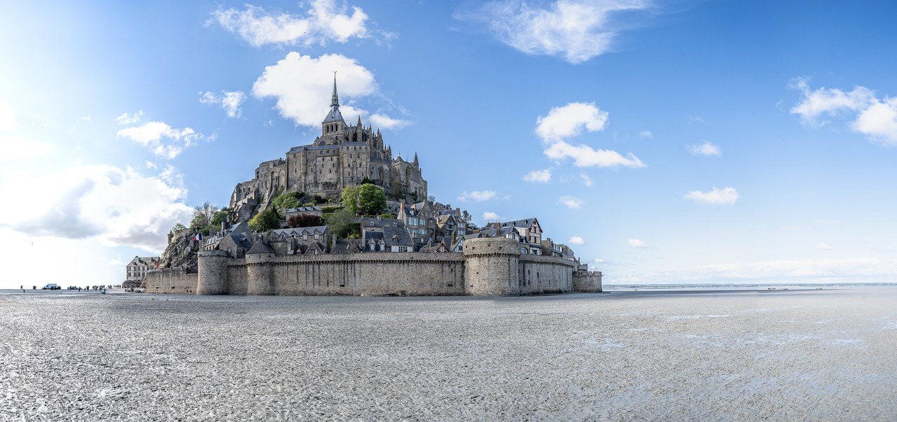 Mont-Saint-Michel seen from the dry strand, the town's curtain wall and its round towers running across the foot of the rock, village roofs stacked above them, and the abbey church and spire on the summit. Mont-Saint-Michel seen from the dry strand, the town's curtain wall and its round towers running across the foot of the rock, village roofs stacked above them, and the abbey church and spire on the summit.