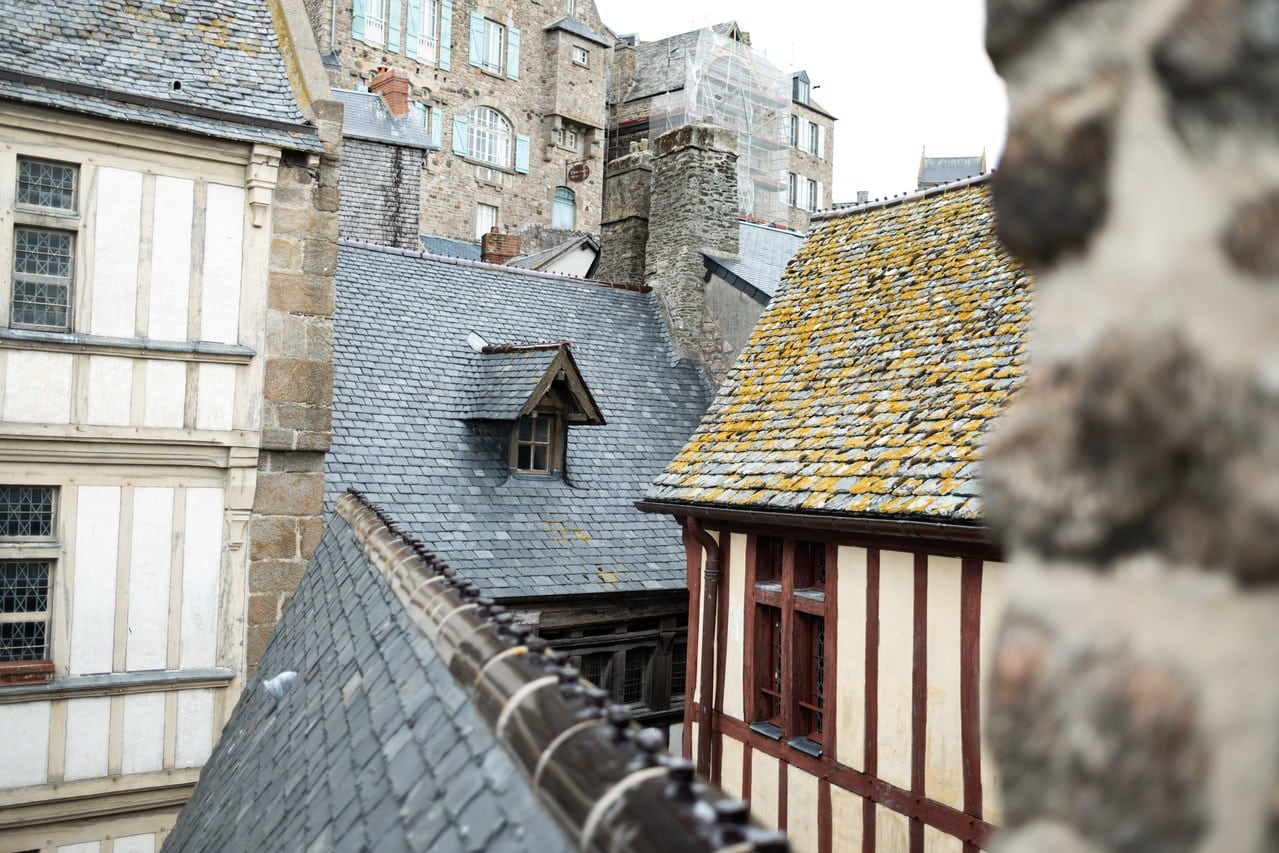 Village roofs at Mont-Saint-Michel seen from the wall-walk: slate and lichen-covered tiles, half-timbered gables and stone chimneys, with scaffolding standing against a building higher up the rock. Village roofs at Mont-Saint-Michel seen from the wall-walk: slate and lichen-covered tiles, half-timbered gables and stone chimneys, with scaffolding standing against a building higher up the rock.