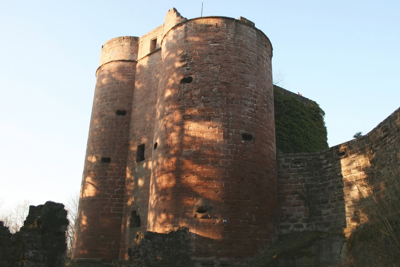 Two broad round towers of red sandstone standing side by side above a rocky slope at Burg Neudahn, their curved faces pierced by small round and slot-shaped openings, with a ruined wall running away to the right. Two broad round towers of red sandstone standing side by side above a rocky slope at Burg Neudahn, their curved faces pierced by small round and slot-shaped openings, with a ruined wall running away to the right.