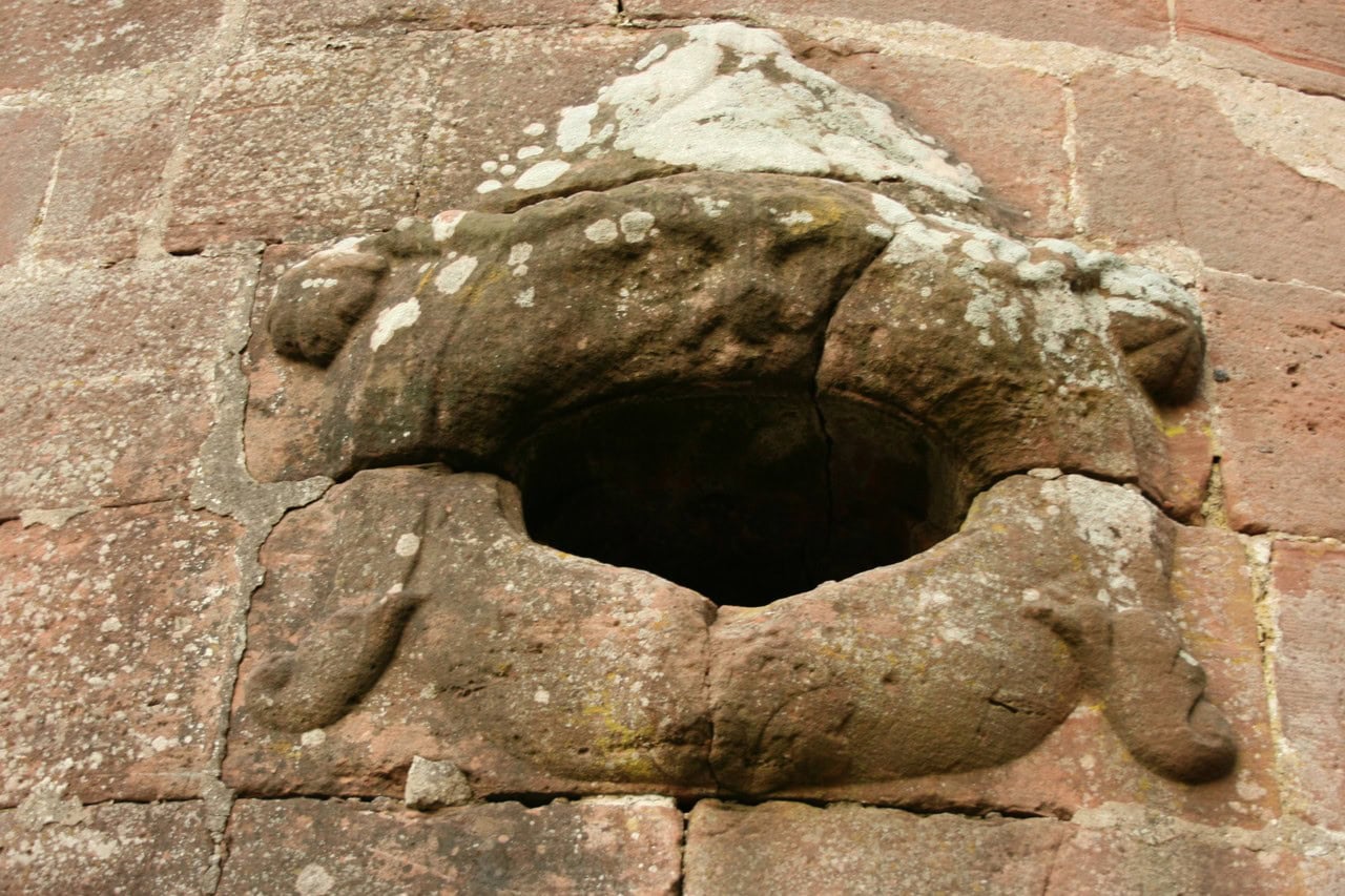 A round gun opening cut through a red sandstone wall at Burg Neudahn, framed by a weathered carved surround with lobed forms above and below the hole. A round gun opening cut through a red sandstone wall at Burg Neudahn, framed by a weathered carved surround with lobed forms above and below the hole.