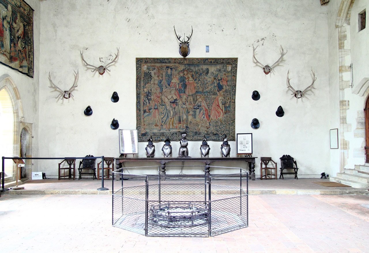 The upper end of a medieval hall: a raised step runs across the far end carrying a long table, a tapestry hangs on the wall above it, and the iron frame of the octagonal floor hearth stands in the foreground. The upper end of a medieval hall: a raised step runs across the far end carrying a long table, a tapestry hangs on the wall above it, and the iron frame of the octagonal floor hearth stands in the foreground.