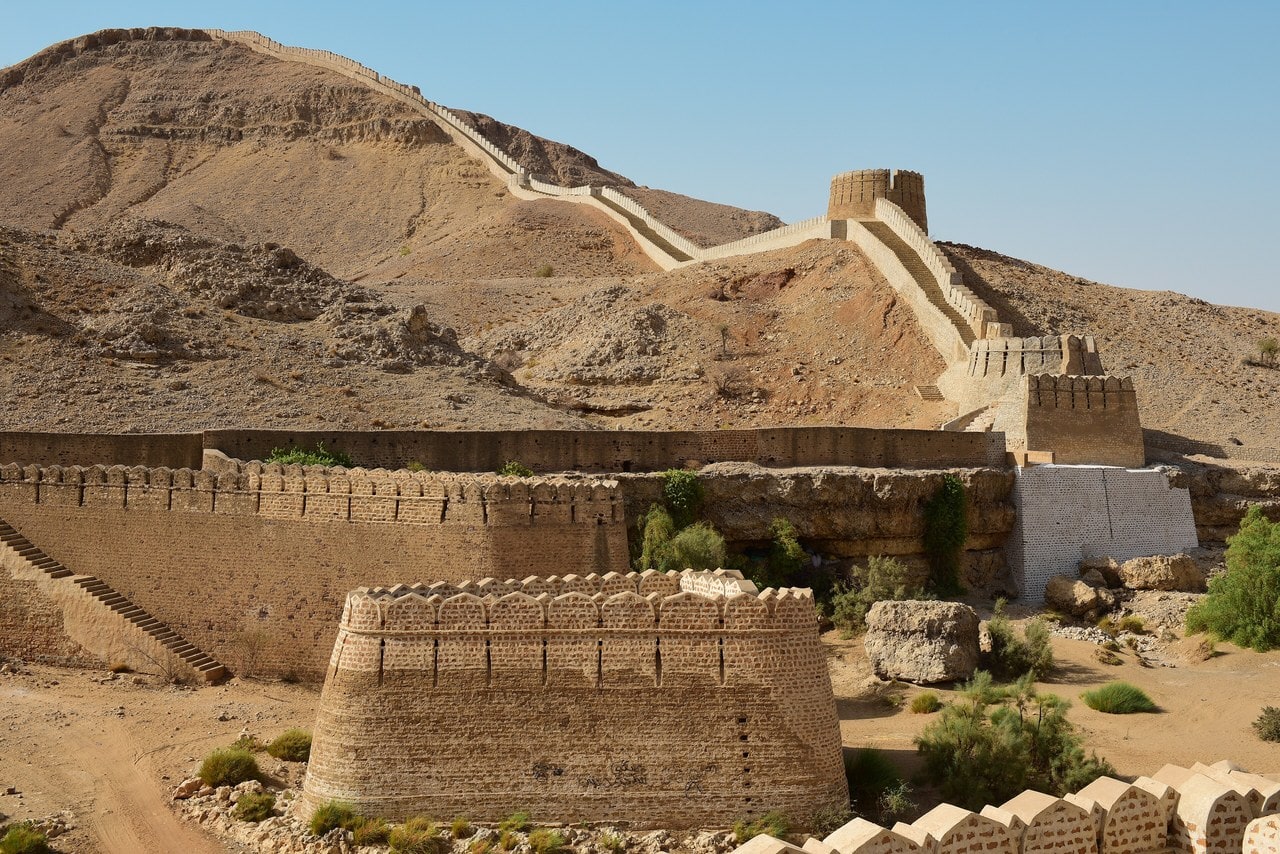 At Ranikot a crenellated wall climbs a bare hillside, passes a round bastion on the slope and continues over the crest; a lower wall with a large round bastion stands in the foreground, and long stretches of the masonry have been rebuilt. At Ranikot a crenellated wall climbs a bare hillside, passes a round bastion on the slope and continues over the crest; a lower wall with a large round bastion stands in the foreground, and long stretches of the masonry have been rebuilt.