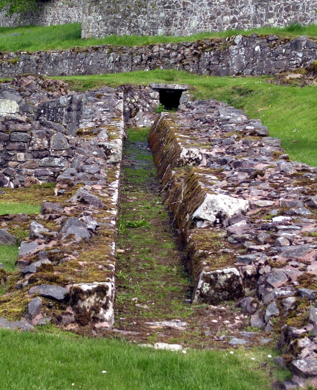 A long straight channel of excavated stonework running away between grass banks, its sides built of rough coursed rubble, with a standing wall behind. A long straight channel of excavated stonework running away between grass banks, its sides built of rough coursed rubble, with a standing wall behind.