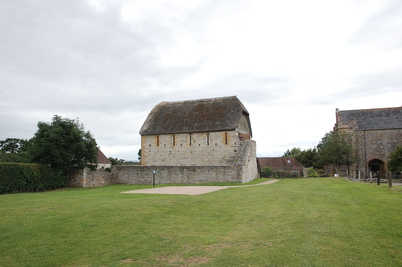 A long two-storey stone building with a thatched roof standing behind a low wall across a mown lawn, with further abbey buildings and trees to either side under a pale sky. A long two-storey stone building with a thatched roof standing behind a low wall across a mown lawn, with further abbey buildings and trees to either side under a pale sky.