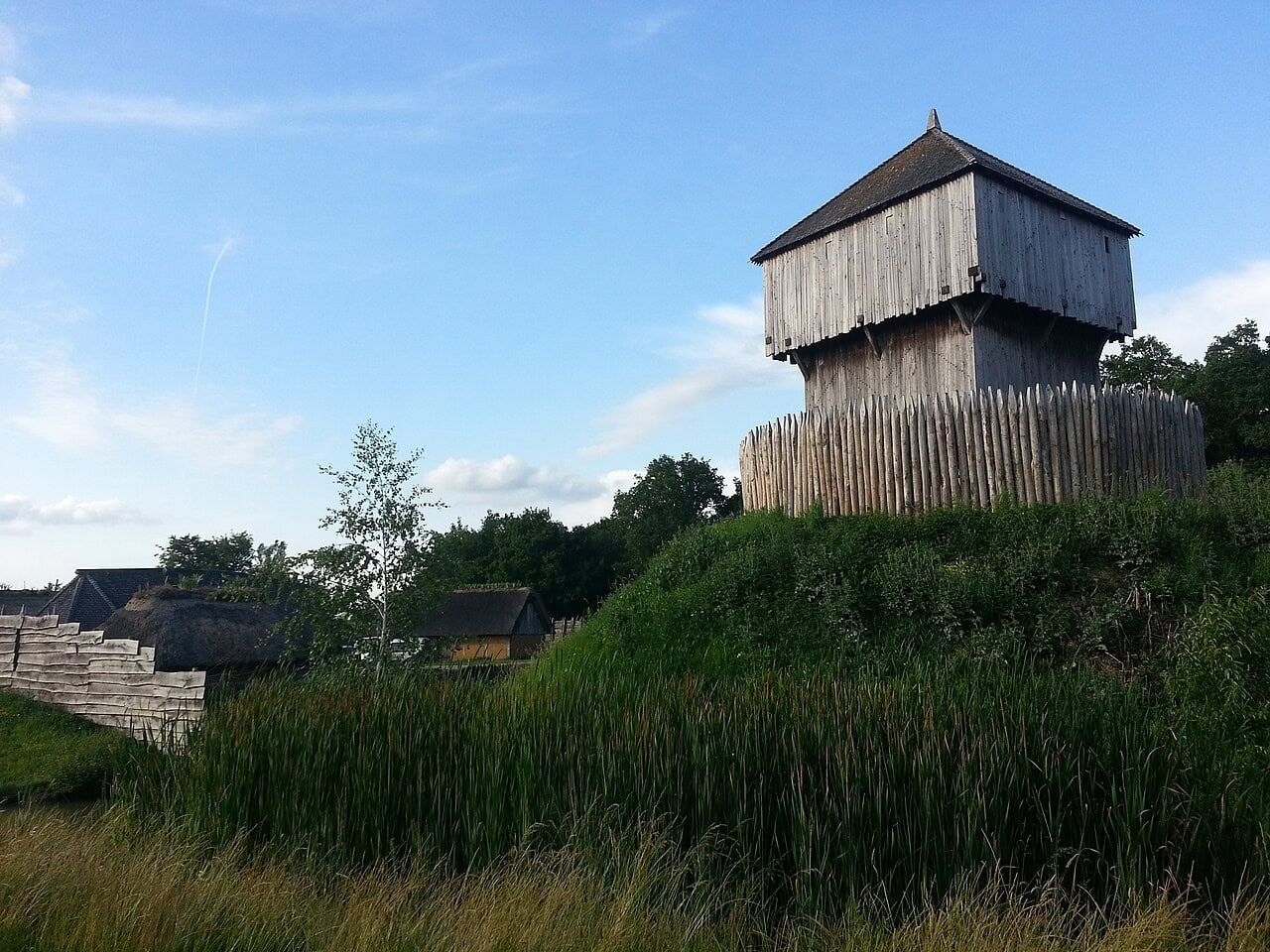 A modern full-scale reconstruction of a timber castle: a boarded tower with an overhanging upper stage and a shingled pyramidal roof stands on the crown of a grassed mound ringed by a palisade of sharpened stakes, with thatched and turf-roofed buildings behind a plank fence on lower ground to the left, and rough grass in the foreground. A modern full-scale reconstruction of a timber castle: a boarded tower with an overhanging upper stage and a shingled pyramidal roof stands on the crown of a grassed mound ringed by a palisade of sharpened stakes, with thatched and turf-roofed buildings behind a plank fence on lower ground to the left, and rough grass in the foreground.