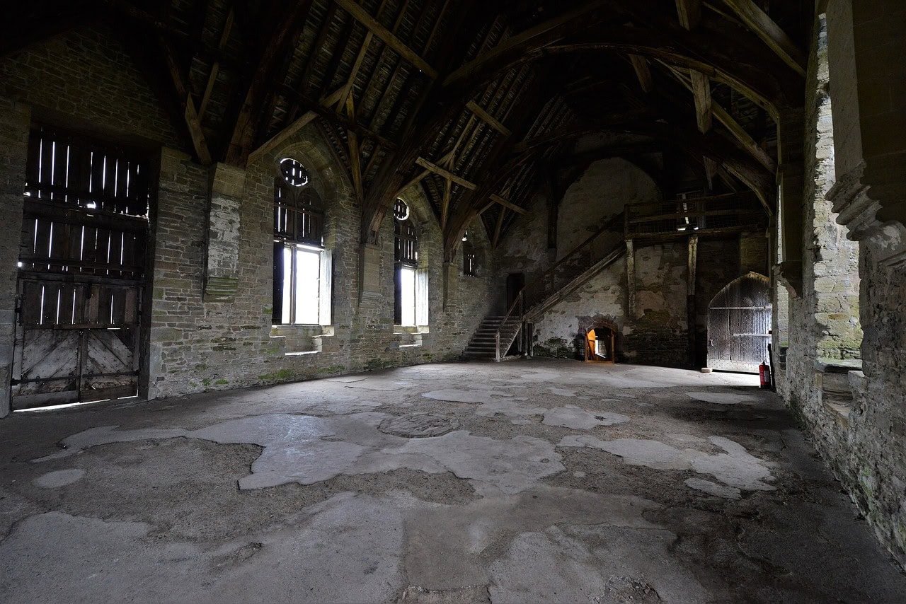 A bare stone hall with a worn floor and no furniture, lit by tall traceried windows in the side wall, its cruck roof open to the rafters overhead and a timber staircase rising against the wall at the far end. A bare stone hall with a worn floor and no furniture, lit by tall traceried windows in the side wall, its cruck roof open to the rafters overhead and a timber staircase rising against the wall at the far end.