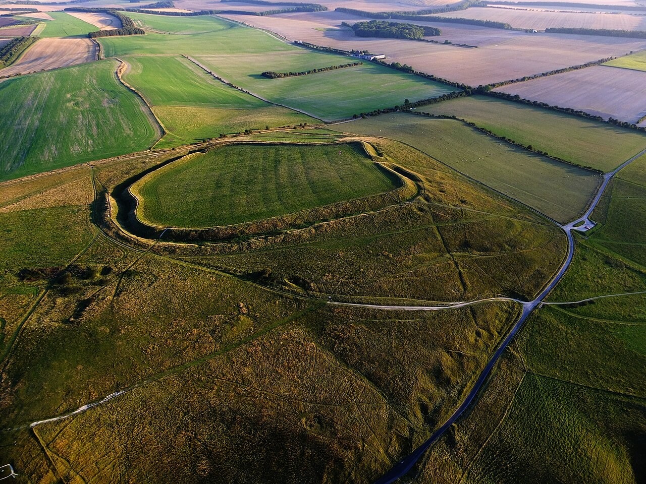 An oblique aerial view of an Iron Age hillfort on open chalk downland: a single grassed rampart and its outer ditch enclose a flat interior, with a gap in the circuit on the far side, and hedged arable fields running to the horizon beyond. An oblique aerial view of an Iron Age hillfort on open chalk downland: a single grassed rampart and its outer ditch enclose a flat interior, with a gap in the circuit on the far side, and hedged arable fields running to the horizon beyond.