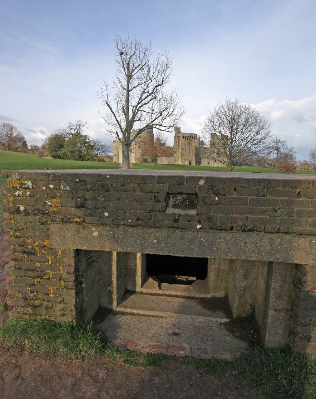 Second World War brick and concrete pillbox on grass with Bodiam Castle visible behind it through bare trees