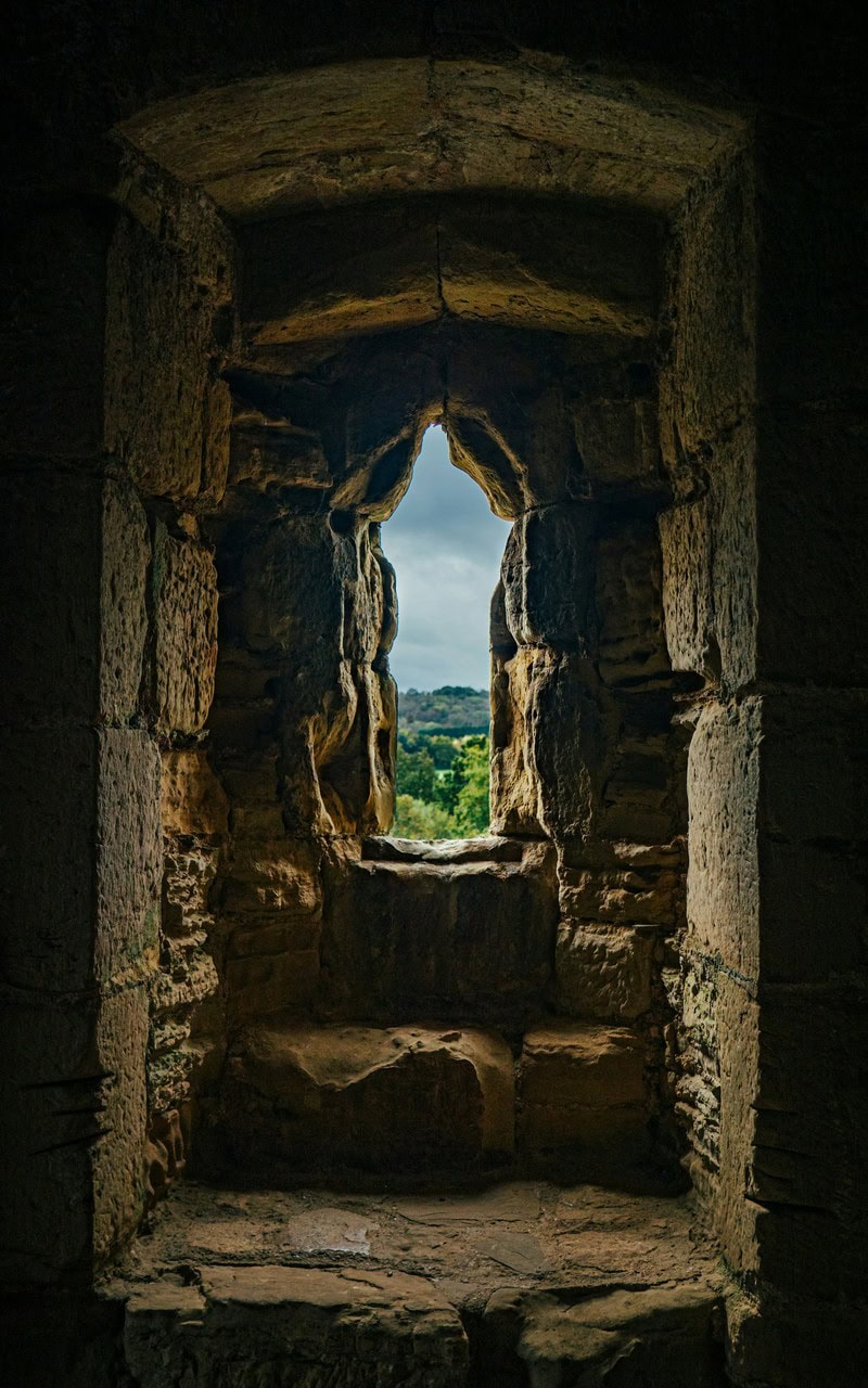 Deep stone embrasure inside Bodiam Castle narrowing to a small pointed opening, trees and a wooded ridge beyond