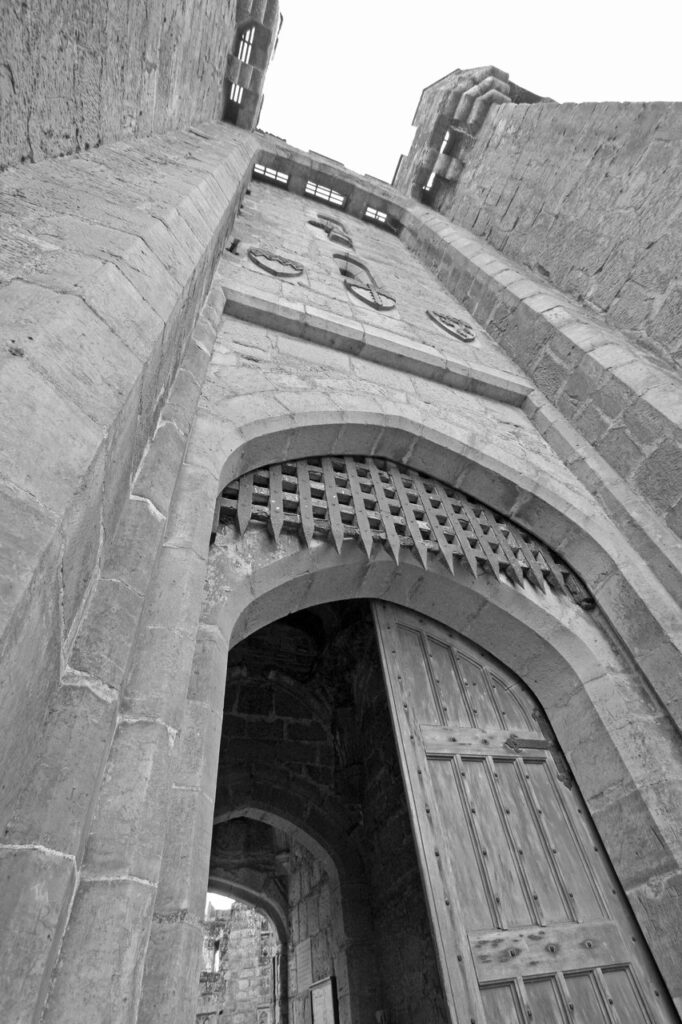 Black-and-white view looking straight up the recessed gatehouse front of Bodiam Castle between its twin towers, three carved shields above the arch, the portcullis raised and a timber door open below