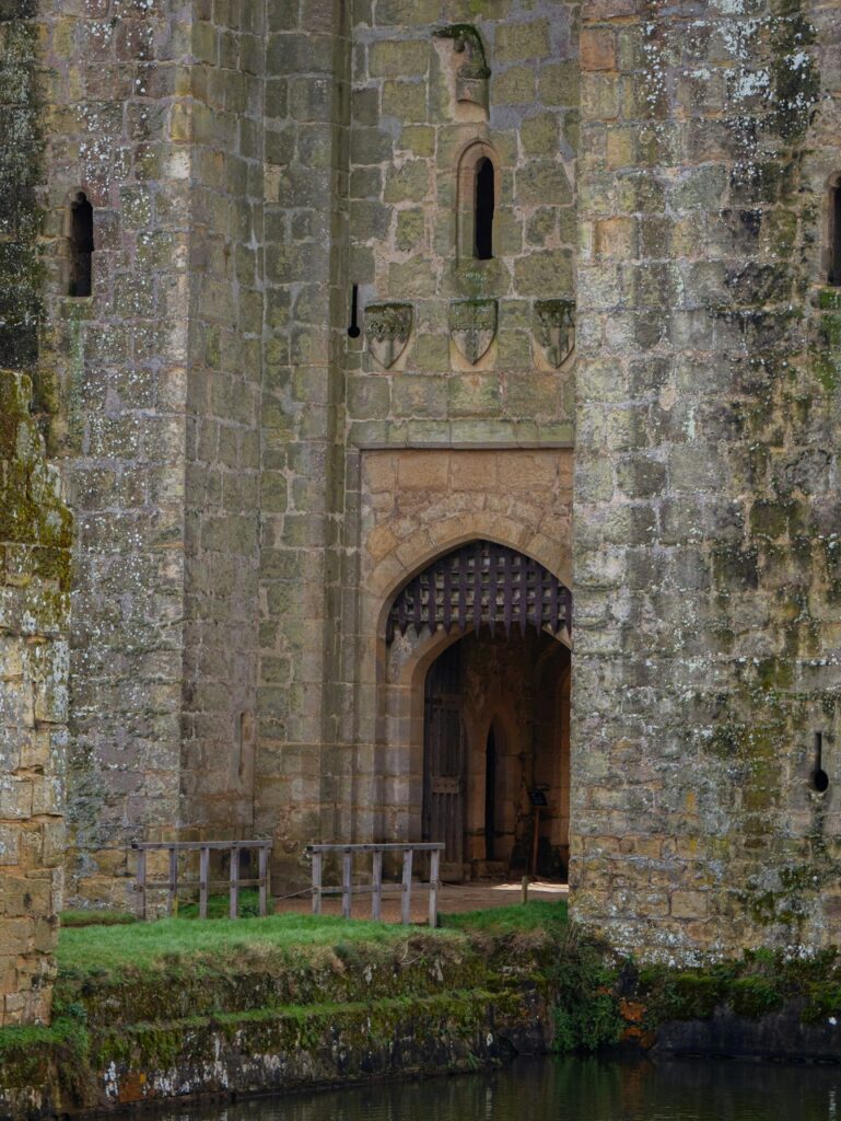 Arched gatehouse entrance at Bodiam Castle seen across the moat, the oak portcullis raised in the opening and three carved shields above