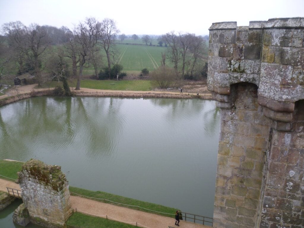 View over the north arm of Bodiam Castle's moat from a tower top, the machicolated gatehouse parapet at right and the barbican ruin on the causeway below