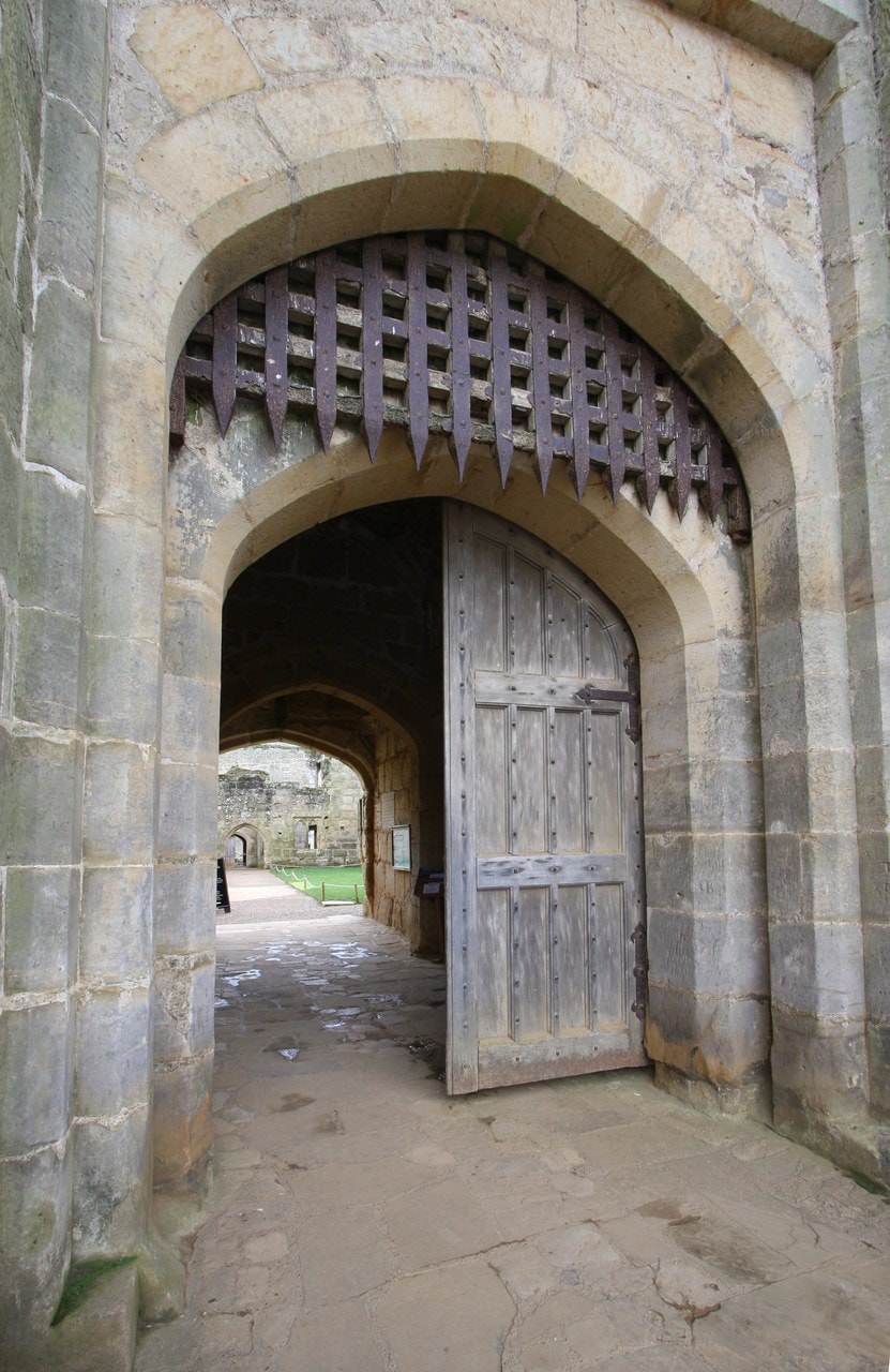 Oak portcullis, iron-clad in its lower part, raised in the stone gate arch of Bodiam Castle, a timber door open on the passage and courtyard beyond