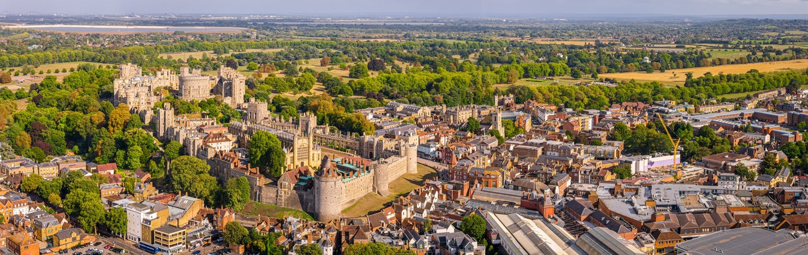 Aerial panorama of Windsor Castle on its hill at the left, its towers and ranges among trees, with the roofs of the town spreading to the right and wooded country to the horizon
