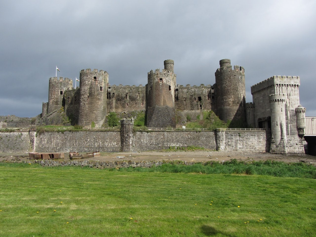 Conwy Castle's south front with the turreted Bakehouse Tower at center, its lower face rebuilt in brown squared stone above a dark, sloping plinth, between round towers and curtain walls, with a pale castellated railway-bridge portal at right.