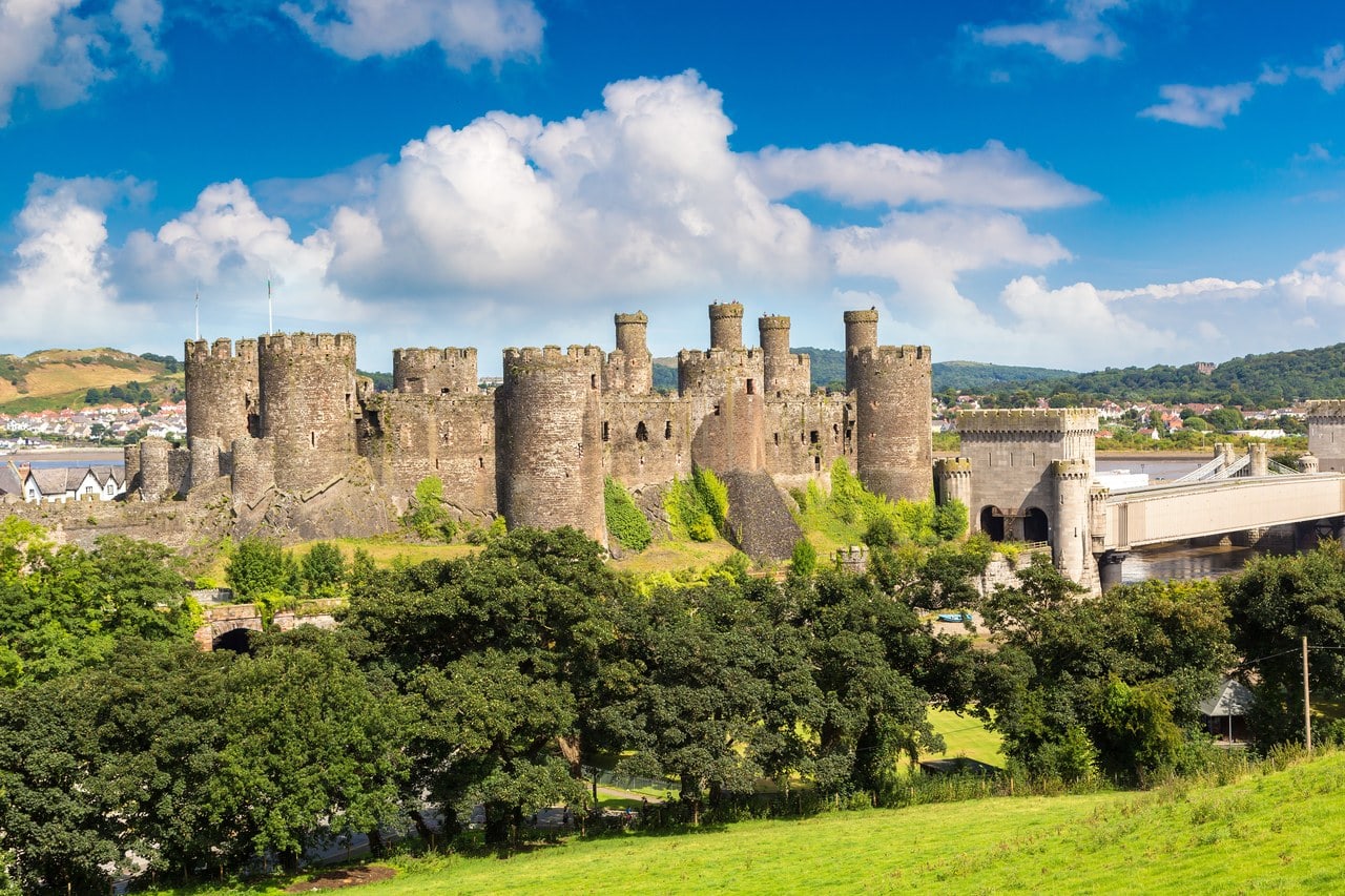 Conwy Castle's long curtain wall and its towers seen across trees from a grassy rise under a blue sky, with the battlemented portal of the tubular railway bridge at the right.