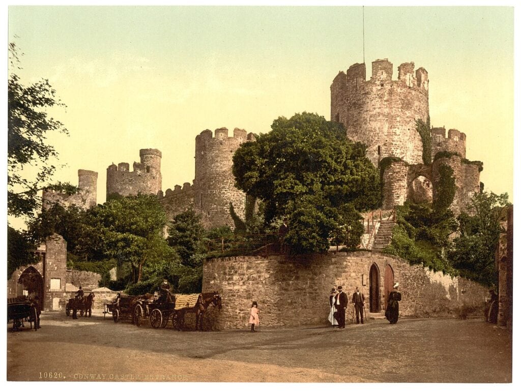 A late nineteenth-century colored photograph of a castle tower and walls above a street with horse-drawn carriages and people