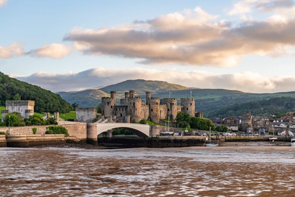 Conwy Castle and the road bridge seen across estuary mud at low tide, hills behind under evening cloud