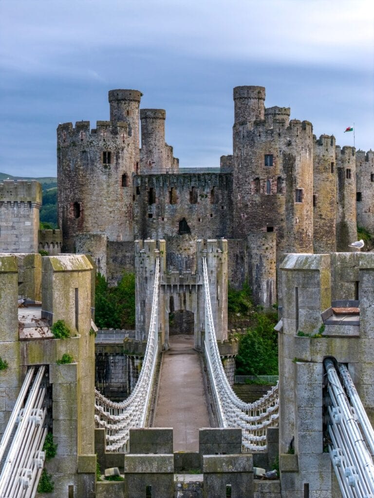An elevated view along Telford's suspension bridge deck, over the tops of the near chain towers, to the castellated bridge towers and Conwy Castle behind them