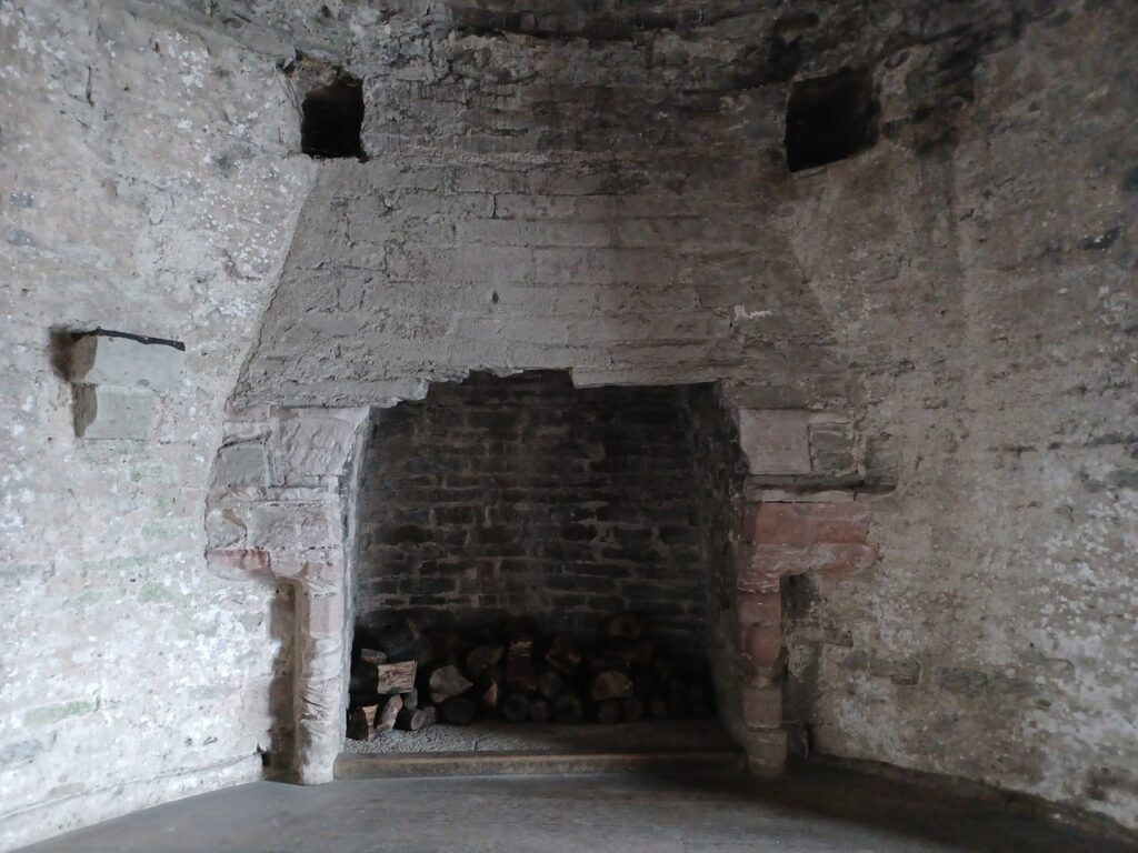 A large stone fireplace with a projecting hood in a whitewashed chamber, logs stacked in the hearth