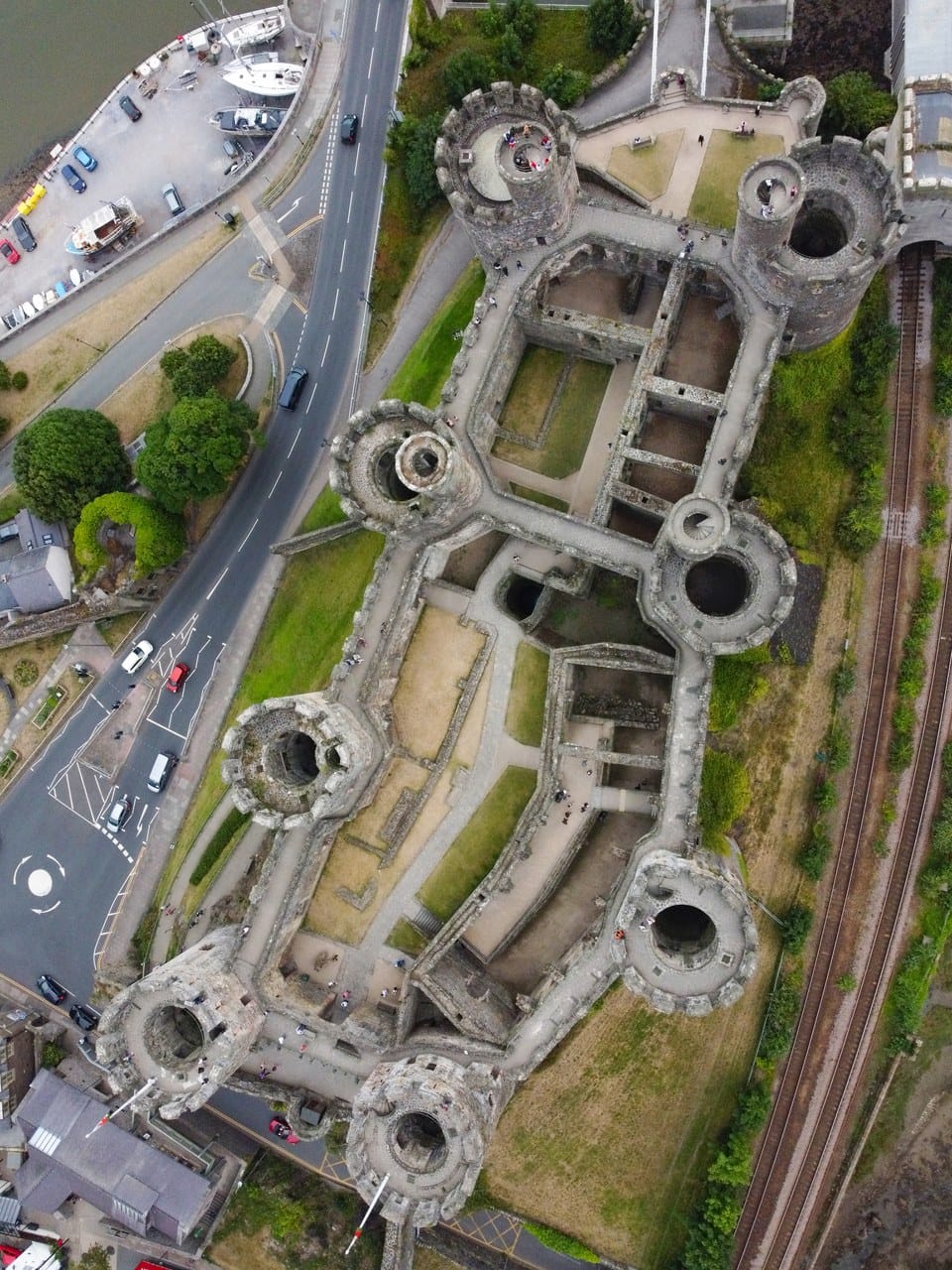 Looking straight down on Conwy Castle: a long narrow walled enclosure with eight towers along its walls and two courtyards inside, a road curving past one side and railway tracks along the other.
