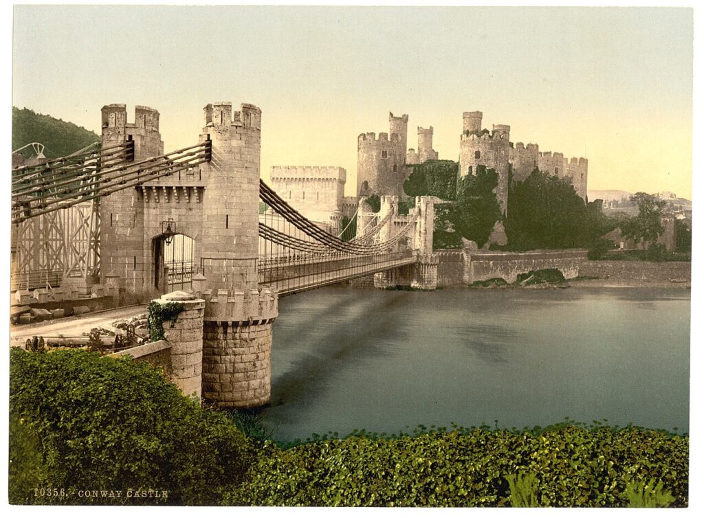 A late nineteenth-century colored photograph of a suspension bridge with castellated towers leading across the water to Conwy Castle