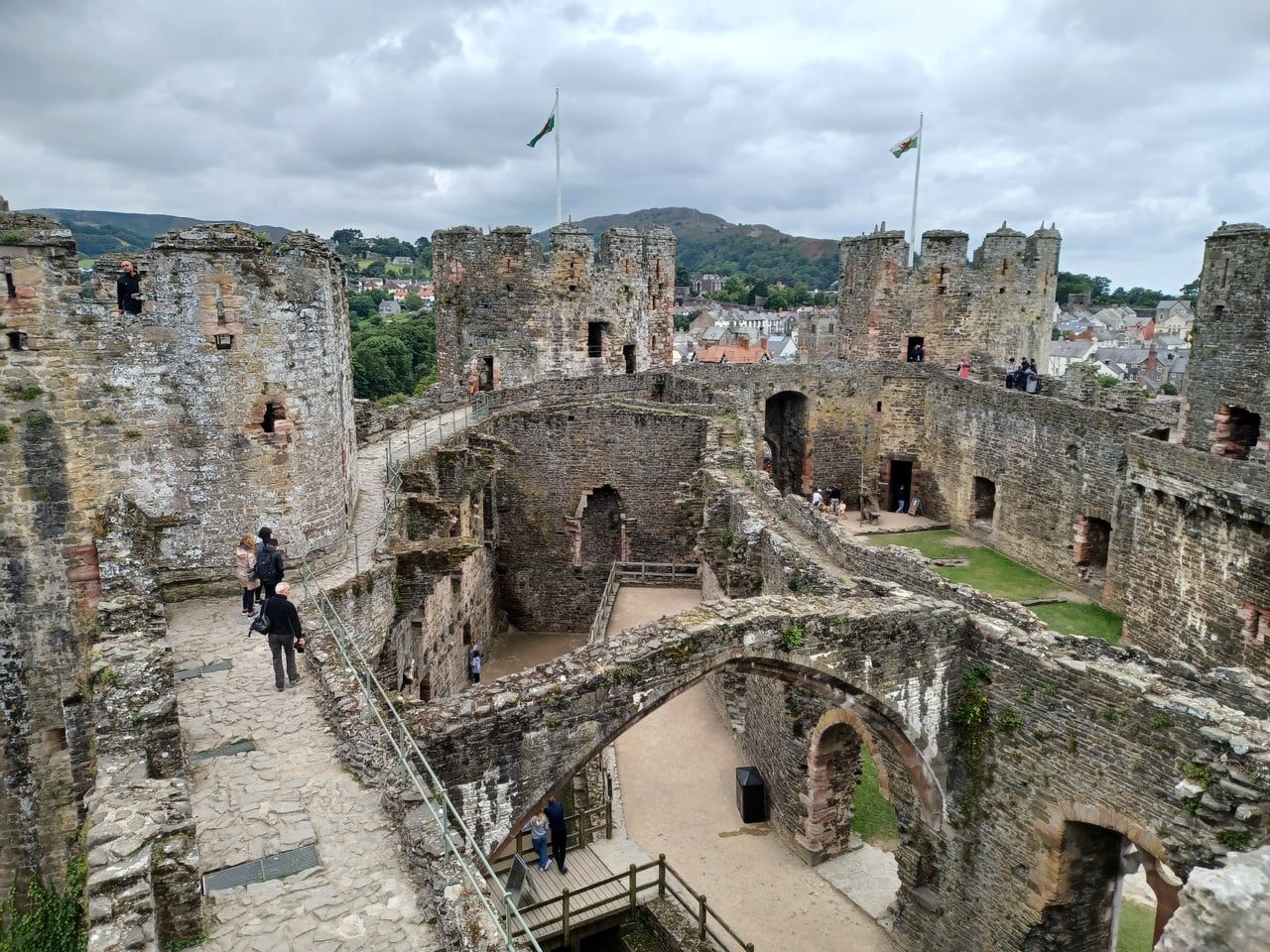 Visitors walking a paved wall-walk along the top of Conwy Castle's curtain, the roofless great-hall range with its one surviving stone arch below and battlemented towers ahead.